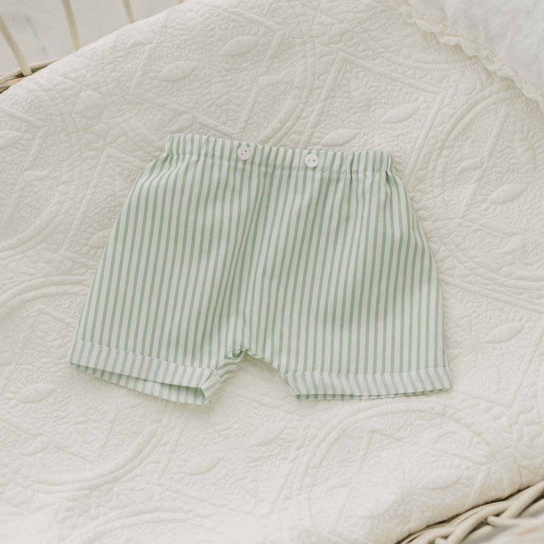 A Theodore Shorts Suit for baby boys in light green and white stripes, featuring two white buttons at the waistband, is laid out on a white, intricately patterned quilt.