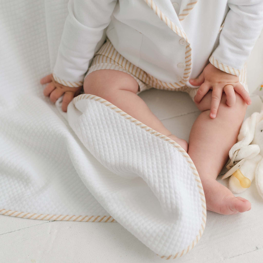 A baby dressed in a white outfit with beige stripes is sitting on a white surface. The baby's legs and hands are visible, and the baby is partially wrapped in the Theodore Personalized Blanket, which features beige striped borders. A stuffed animal is partially visible beside the baby, making it the perfect personalized baby gift.