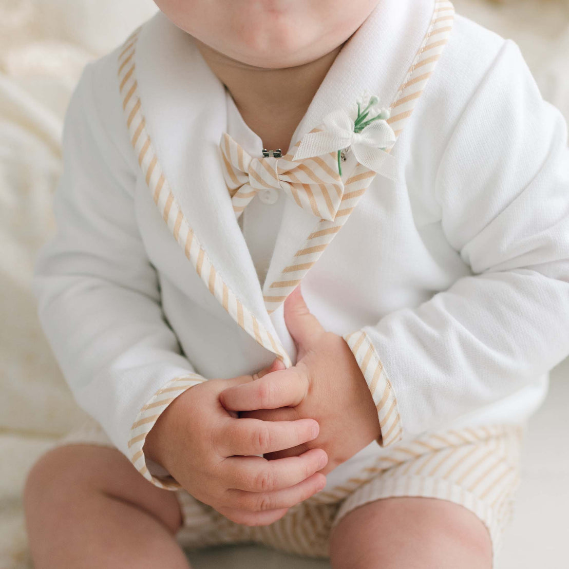 A baby is dressed in a Theodore Shorts Suit, crafted from French Terry cotton with beige striped accents. The handmade outfit includes a bow tie, a jacket with striped trim, and matching shorts. The baby is holding the lapel of the jacket with both hands and has a small flower pinned to it.