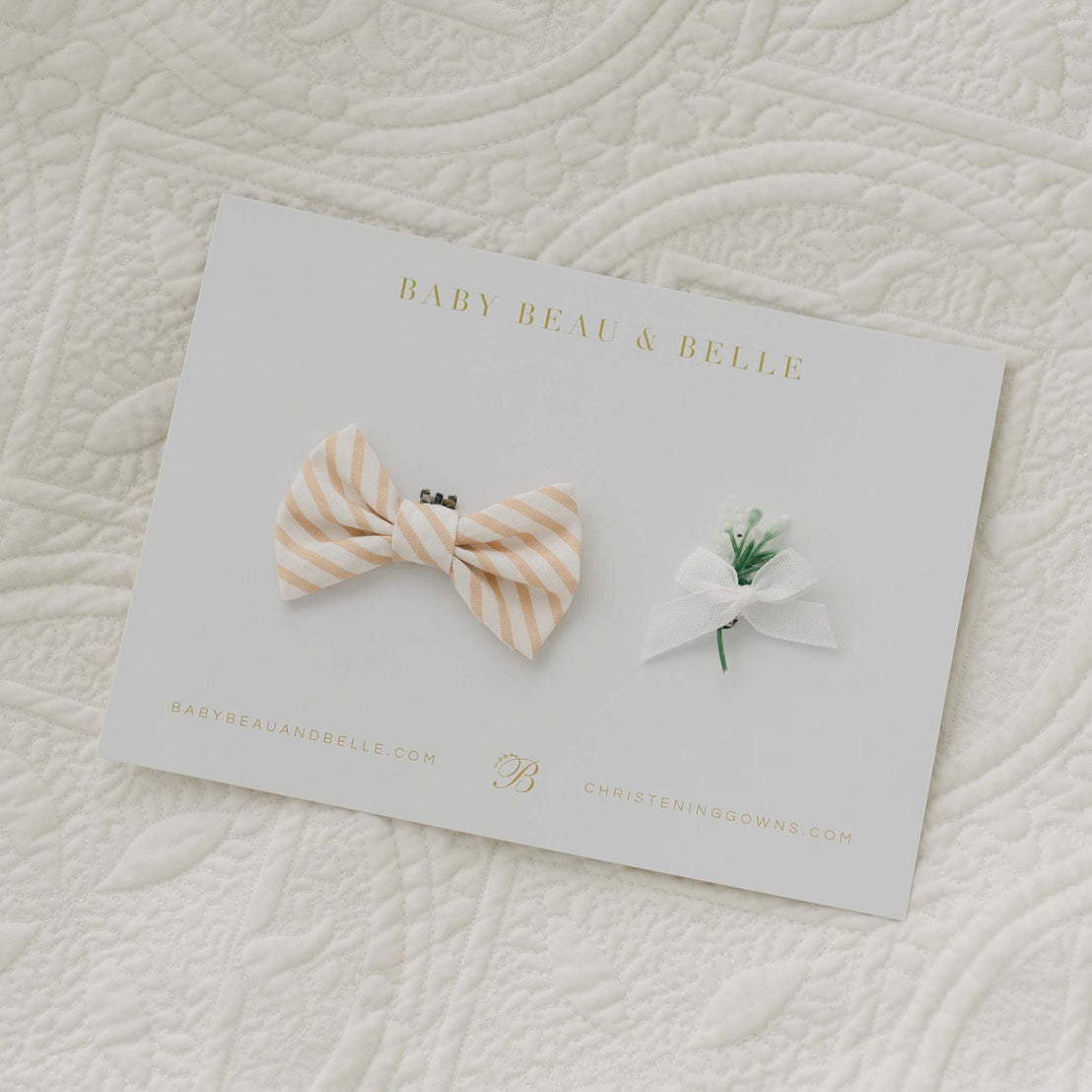 A card labeled "Theodore Bow Tie & Boutonniere" showcases a handmade bow tie crafted from beige and white striped cotton, accompanied by a small white flower with a ribbon. The card is placed on quilted white fabric, with "www.babybeaubelle.com" and "christeninggowns.com" printed at the bottom.