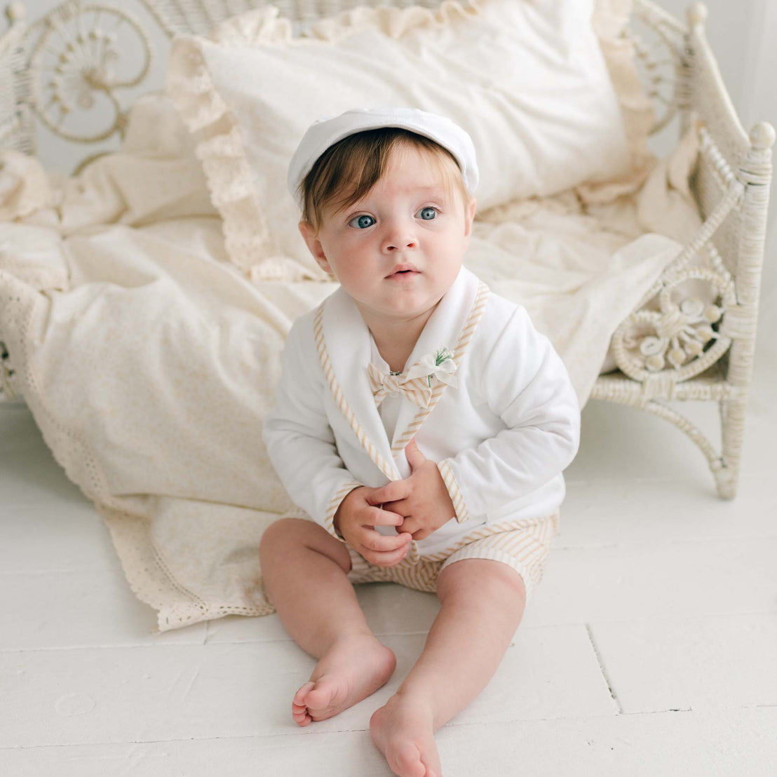 A baby with light hair and blue eyes sits on a white floor, dressed in the Theodore Shorts Suit featuring beige stripes, a matching hat, and a small boutonniere. Behind the baby is a vintage-style white wicker crib with cream-colored bedding. The baby looks slightly upward.