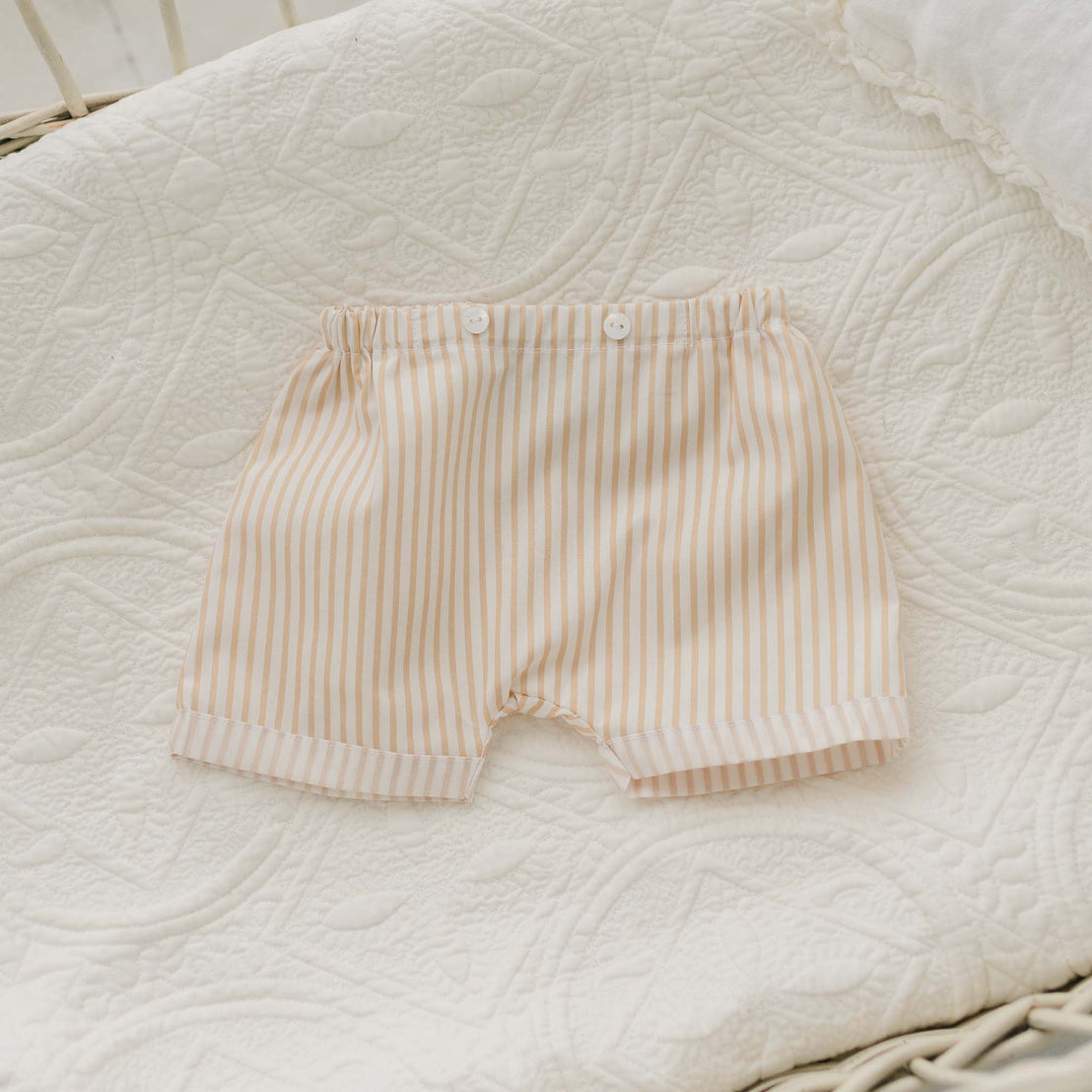 A Theodore Shorts Suit, featuring a pair of beige and white striped baby boy shorts with an elastic waistband and button detailing at the front, laid flat on a white, textured quilt.