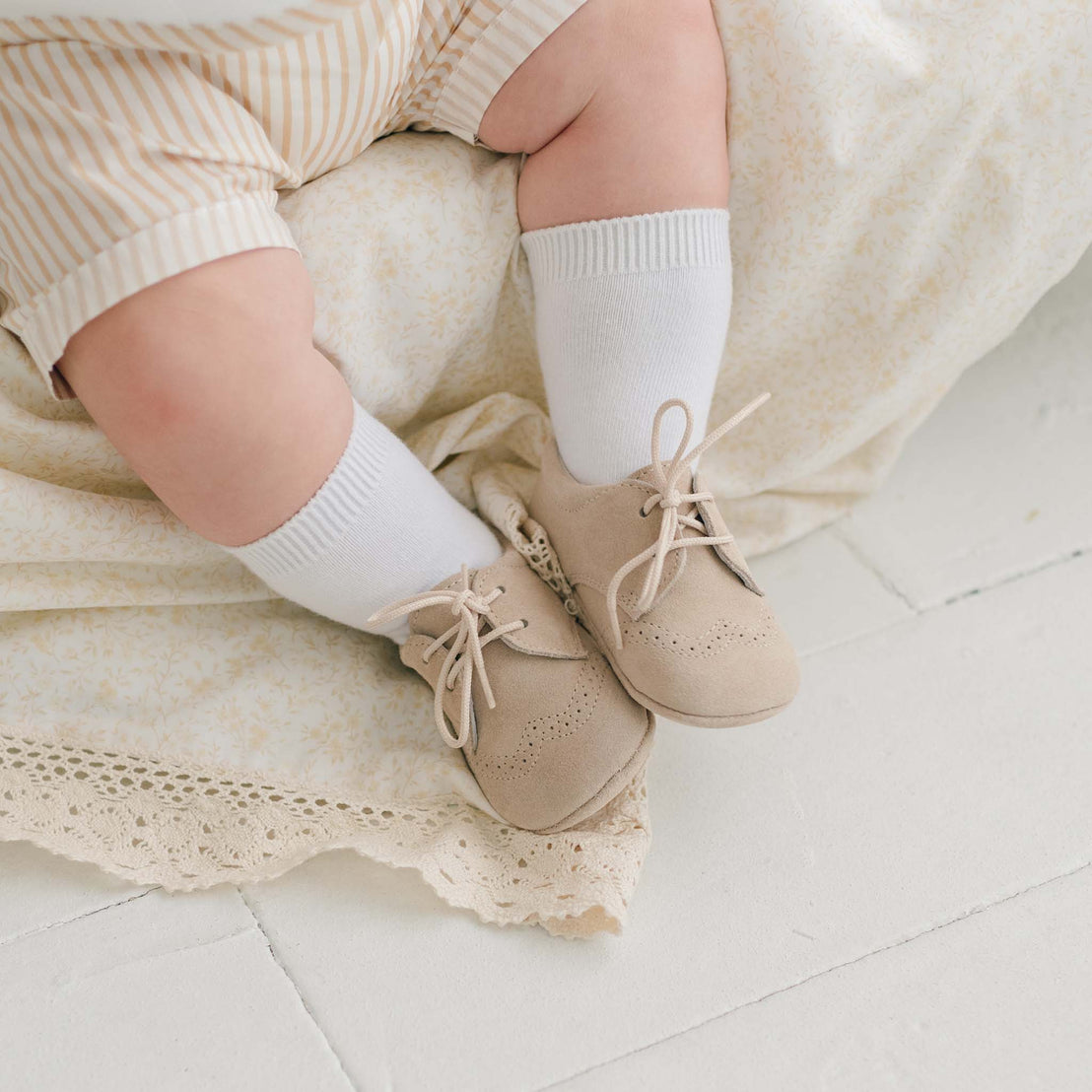 A close-up of a baby's legs wearing light-colored striped shorts, white socks, and Theodore Suede Shoes by Baby Beau & Belle. The baby is sitting on a light-colored fabric with lace trim on a white floor.