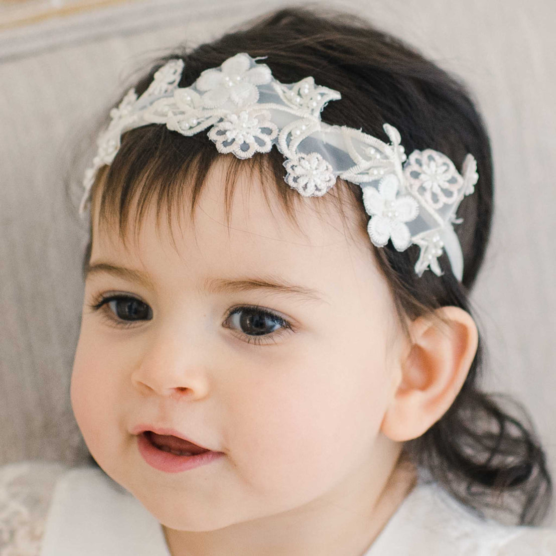 A close-up of a smiling toddler with dark hair wearing the Jessica Beaded Flower Headband, a delicate white and pink floral headband.