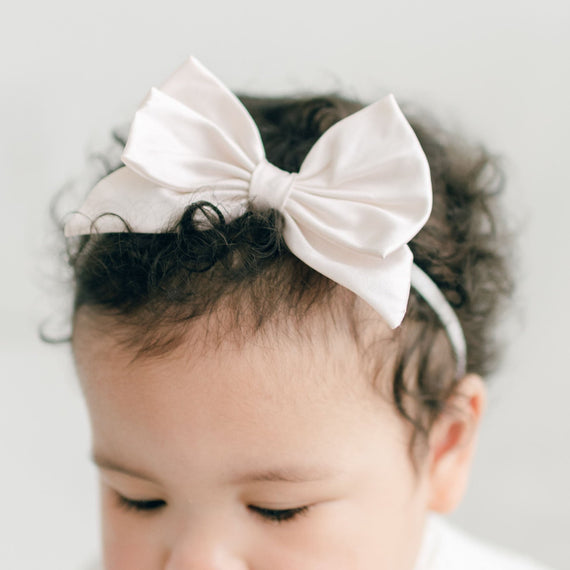 A close-up of a baby with curly hair wearing a Victoria Silk Bow Headband, set against a light gray background. The baby has a thoughtful expression.