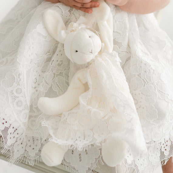 A child in a lace dress holds a Victoria Silly Bunny Buddy | Pacifier Holder adorned with lace and small flowers. The bunny, designed to be baby-safe, doubles as a pacifier holder. The child’s hands and part of the dress are visible, emphasizing the delicate and intricate lace details. The scene exudes a sense of innocence and tenderness.