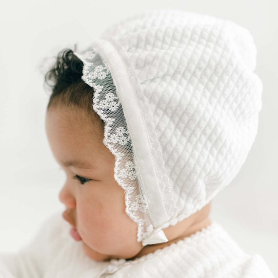 A close-up of a baby wearing a delicate Victoria Quilted Cotton Bonnet adorned with floral lace, focusing thoughtfully off to the side. The baby is dressed in a white outfit, providing a soft, pure appearance.