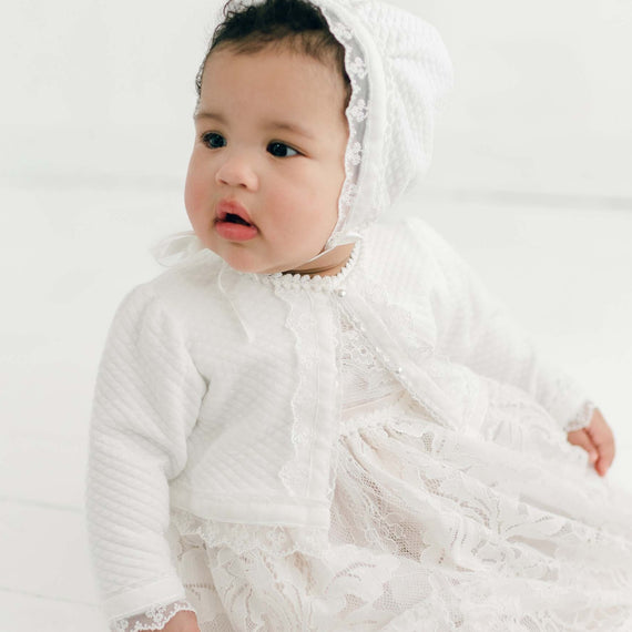 A baby dressed in a Victoria Quilted Cotton Sweater gown and bonnet looks off to the side with a curious expression in a bright, all-white studio setting.