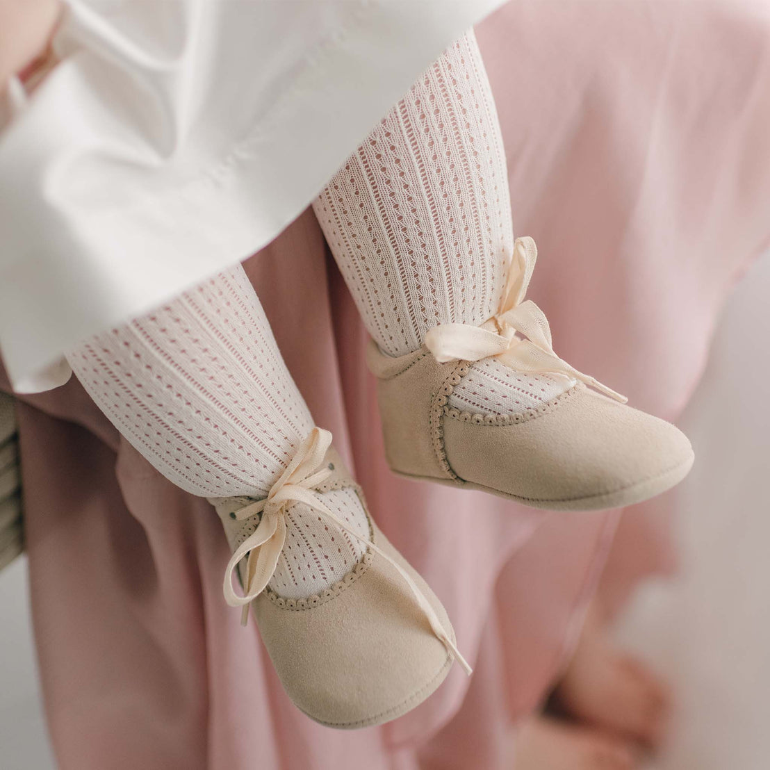 Close-up of a baby's legs wearing cream-colored, knit tights and Tan Suede Tie Mary Janes. The baby is seated on someone's lap, partially visible, who is wearing a pale pink skirt. The background is softly blurred.