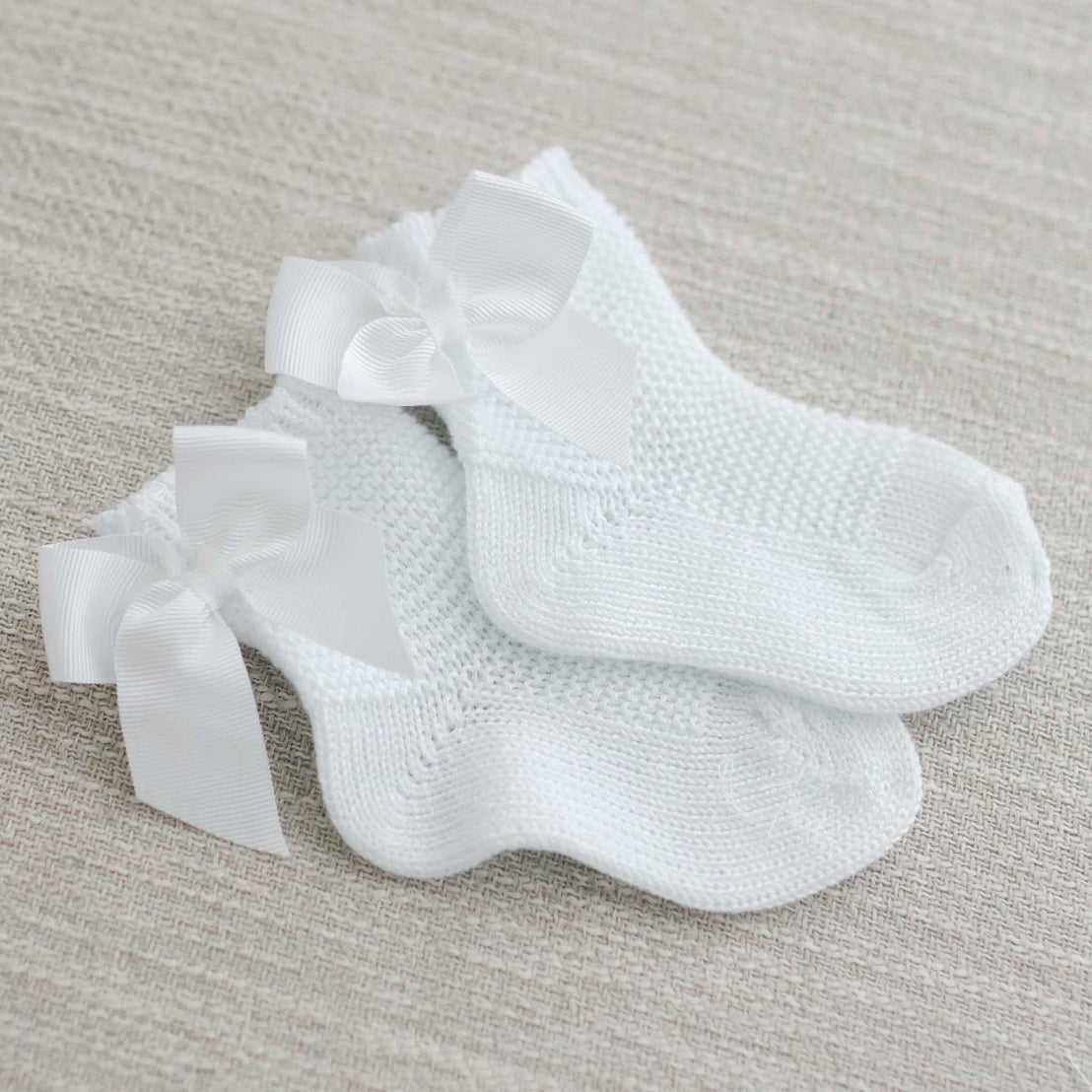 A pair of Garter Stitch Socks with Bow in white neatly placed on a textured beige fabric background.