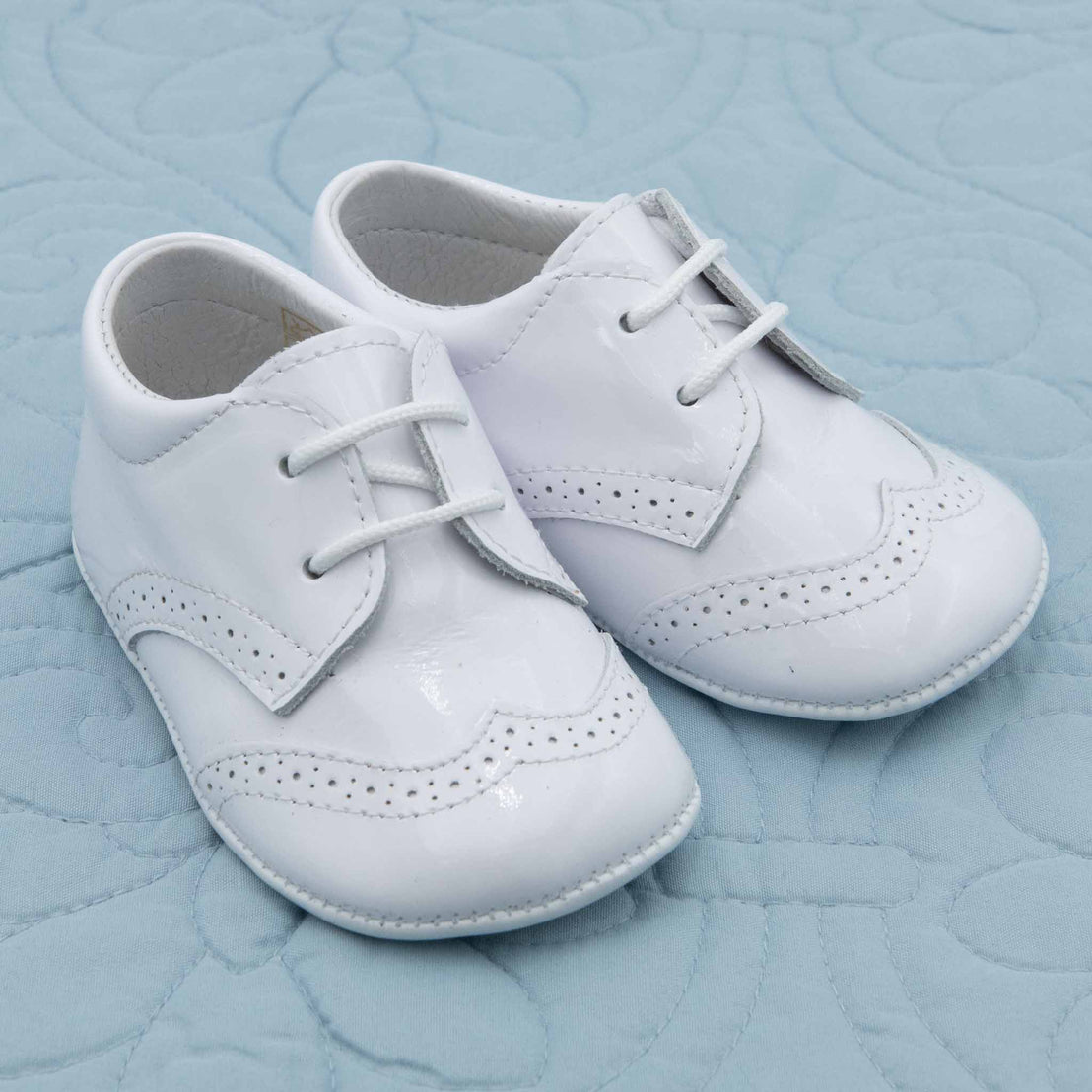 A pair of White Patent Leather Shoes placed on a light blue quilted surface.  The shoes feature decorative perforations and stitching.