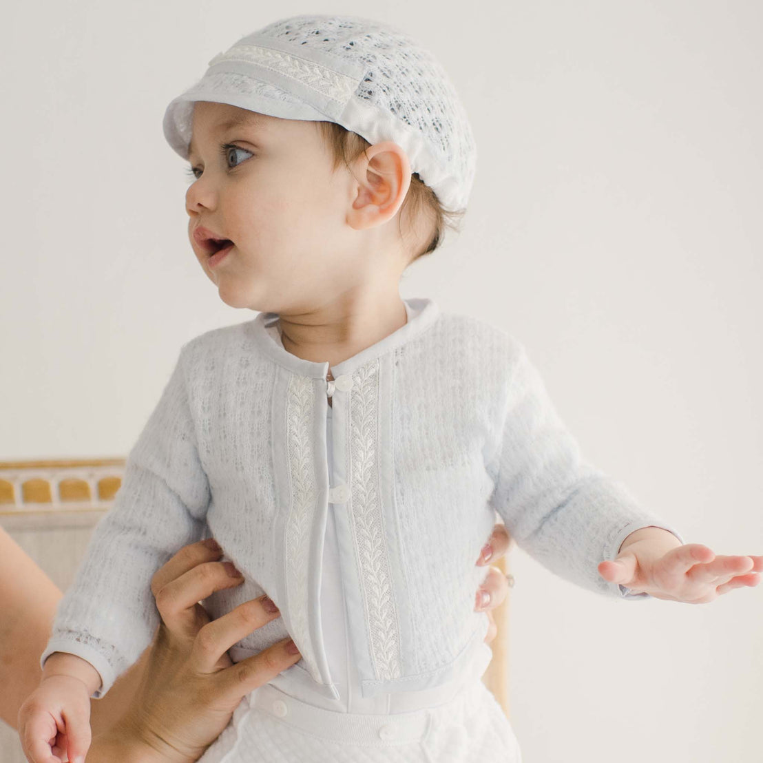 A baby wearing a light-colored hat and a white outfit with intricate detailing is being supported by an unseen adult's hands. The baby, looking to the side, also dons a Harrison Blue Knit Sweater. The background is plain and light-colored.