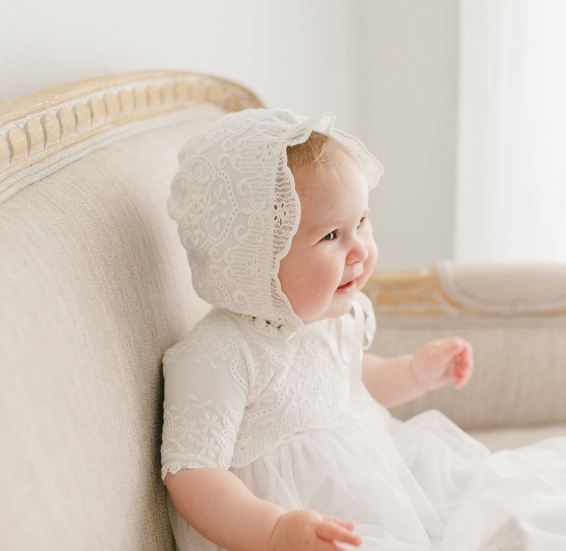 A joyful baby in the Eliza Bonnet and matching Eliza Christening Bonnet sits on a beige sofa, smiling and looking to the side with a bright expression.