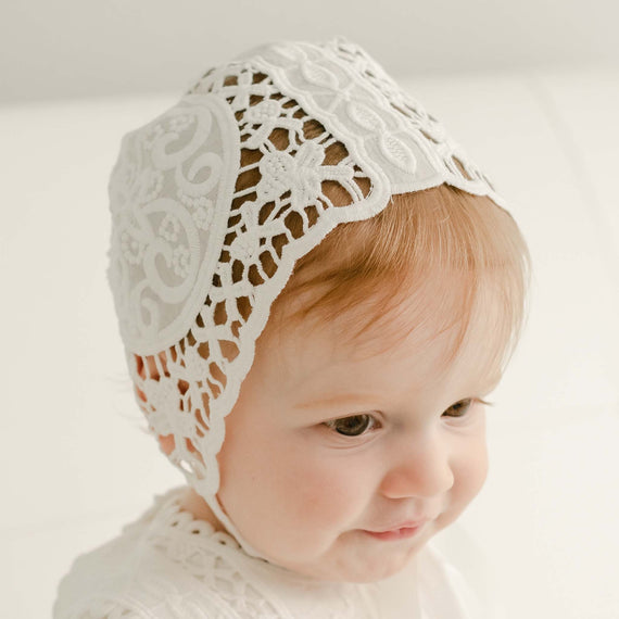 A baby wearing a delicate, intricately designed Adeline Lace Bonnet looks downward with a slight smile. The baby's light hair is partially visible under the bonnet, and the background is softly lit and neutral. A silk ribbon adds an extra touch of charm to this adorable scene.
