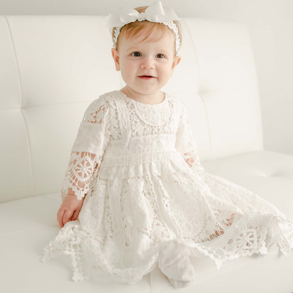 A toddler in the Adeline Lace Dress & Bloomers and matching bow headband, adorned with baby jewelry, sits on a luxury white couch, looking directly at the camera with a soft smile.