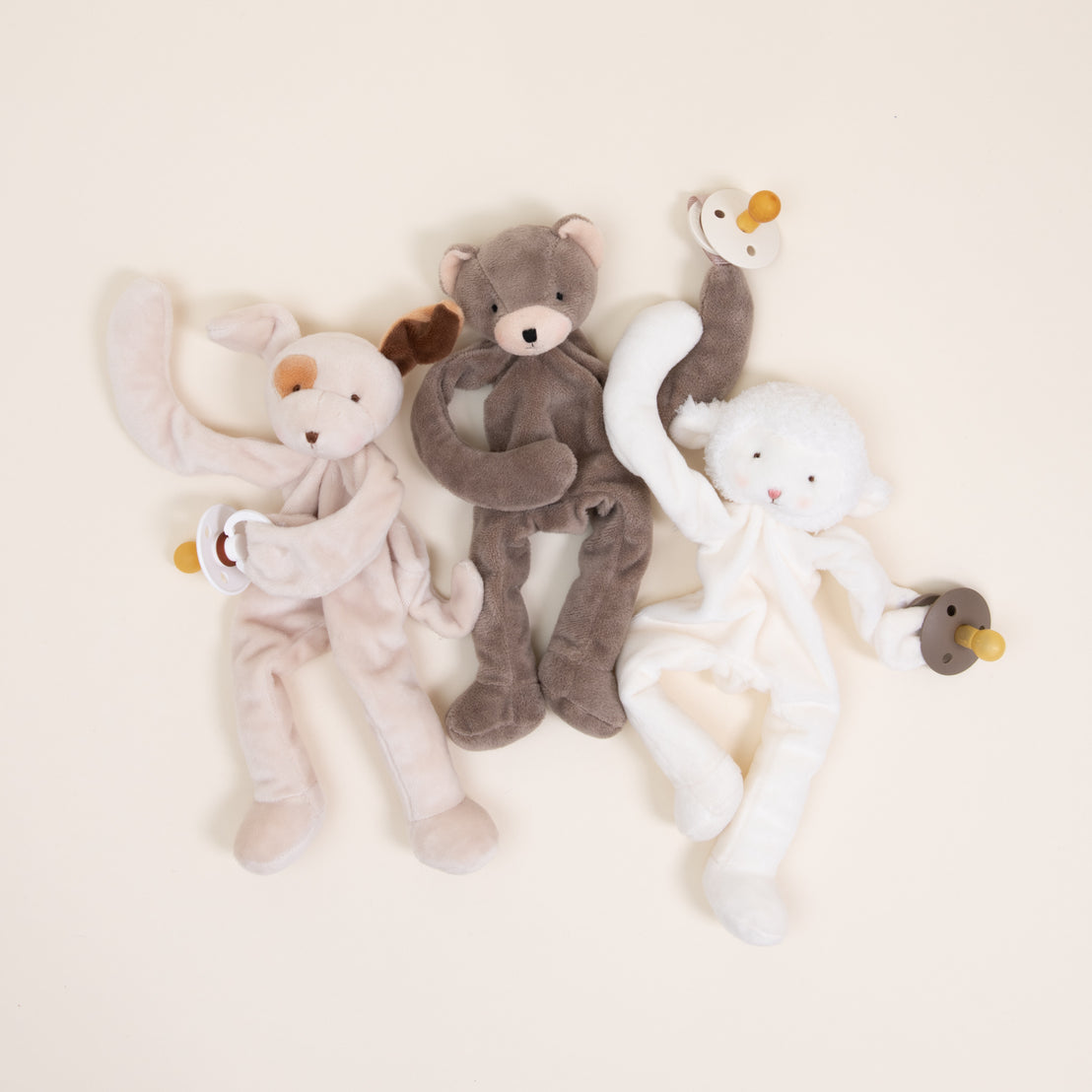 Three Silly Buddy Pacifier Holders, including a rabbit, a bear, and a lamb from Baby Beau & Belle, each holding a small wooden ring with a pacifier clip, arranged side by side on a light beige background.