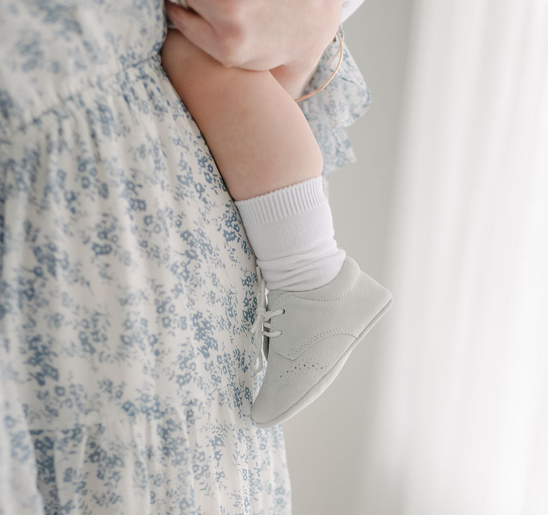 Photo of a baby foot wearing the Miles Suede Shoes. Made with a soft grey suede with detailed edging.