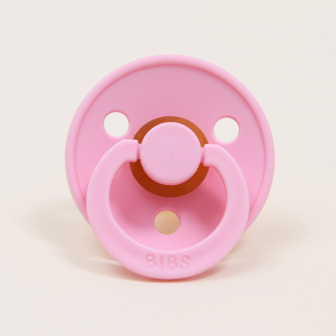 A pink bibs pacifier with three air holes and a brown silicon nipple on a plain, light-colored background.