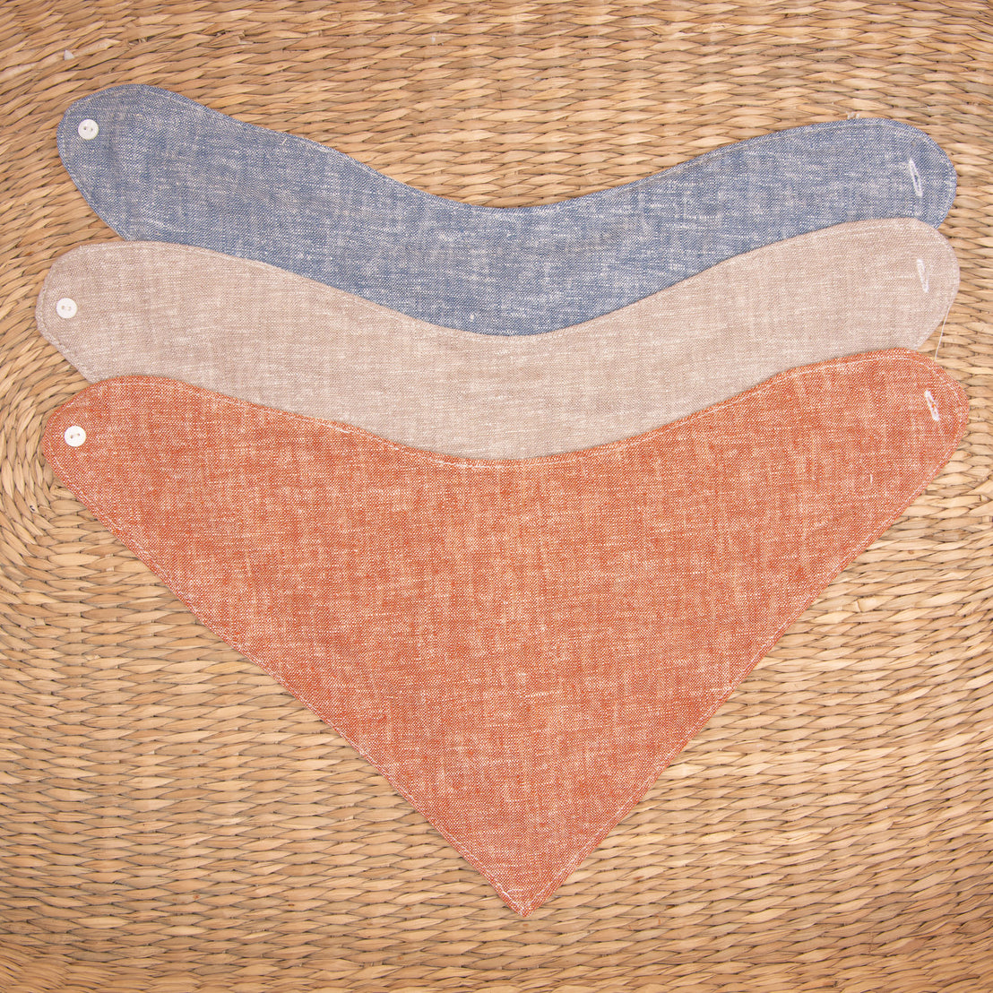 Flat lay photo of all three colors of the Silas Bandana Bib. The bibs are made from linen in three different colors: indigo, sand, and clay.