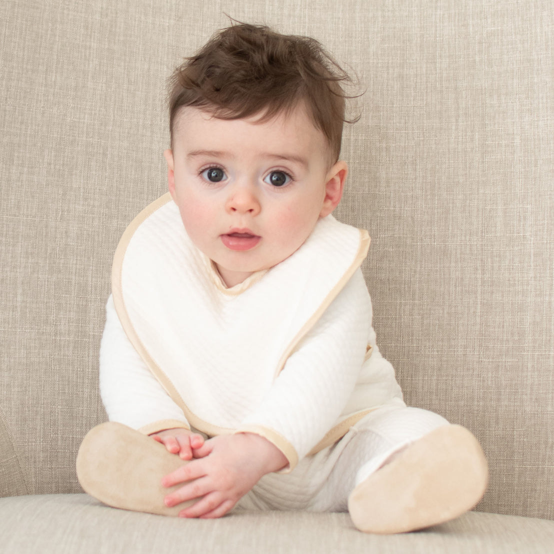 Baby boy sitting on a chair and wearing the Liam Boys Bib crafted from soft quilted cotton in ivory and detailed with a silk champagne trim