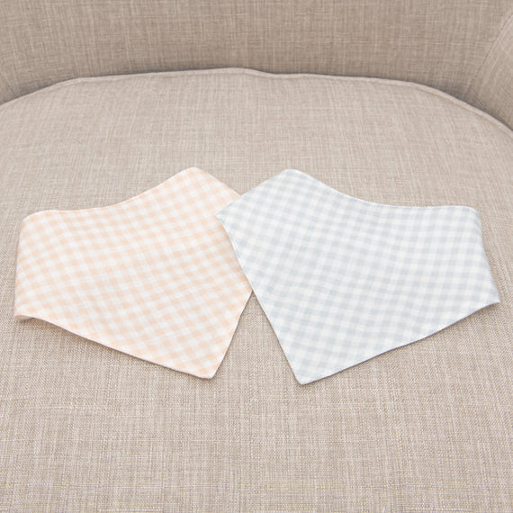 Two Isla Bandana Bibs with checkered patterns, one peach and one light blue, lie on a upholstered beige sofa.