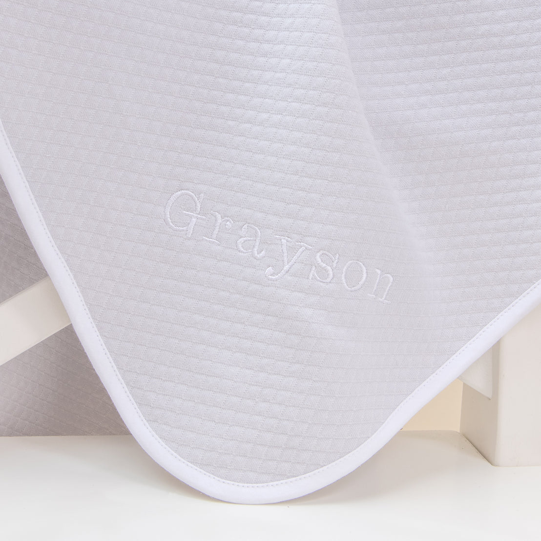 Flat lay photo of the Grayson Personalized Blanket. It is made from quilted textured cotton in grey with a white pima cotton trim. On the corner is the name "Grayson" embroidered in white thread.