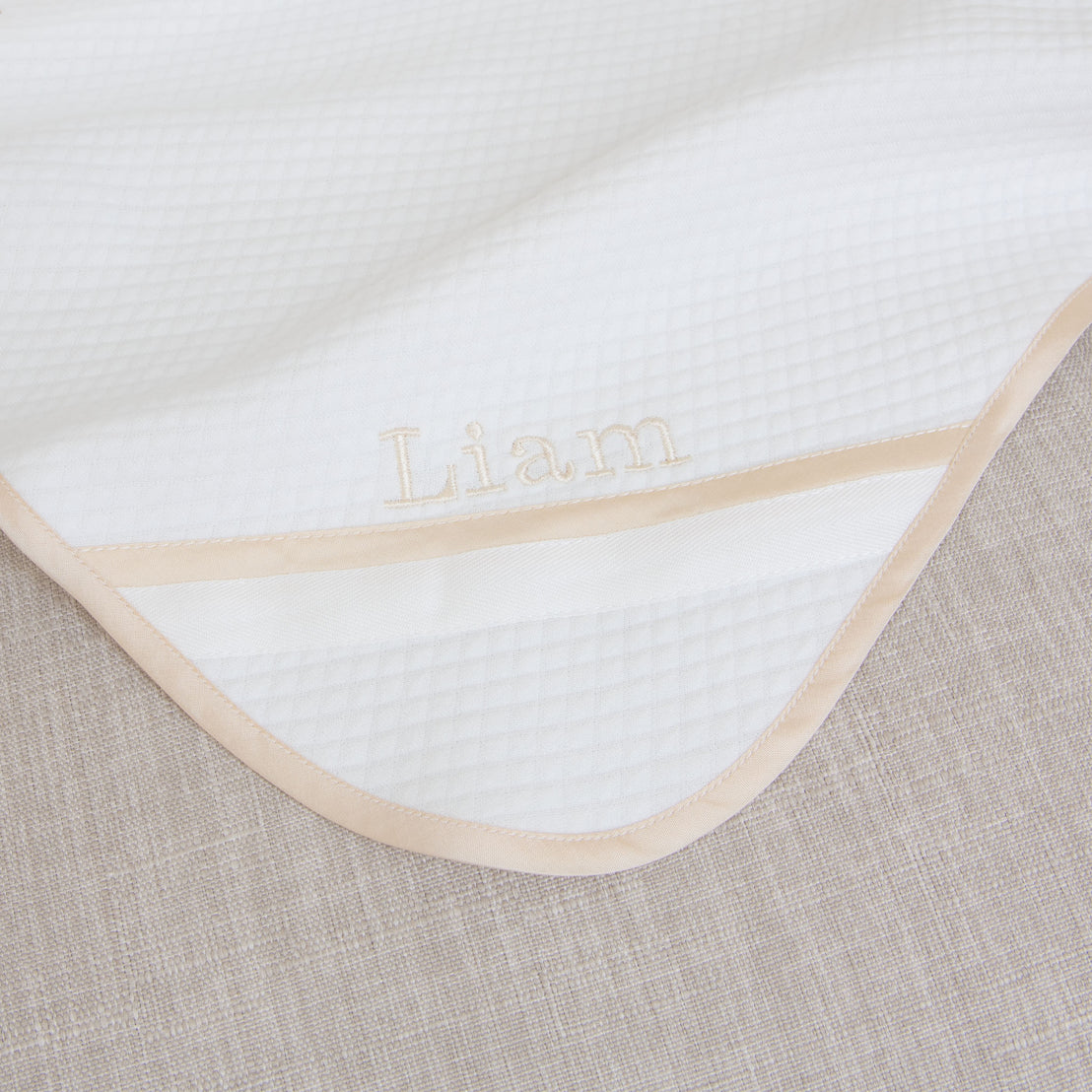 Flat lay of the Liam Personalized Blanket made with a soft ivory quilted cotton and champagne trim. Embroidered on the corner of the blanket is the name "Liam"