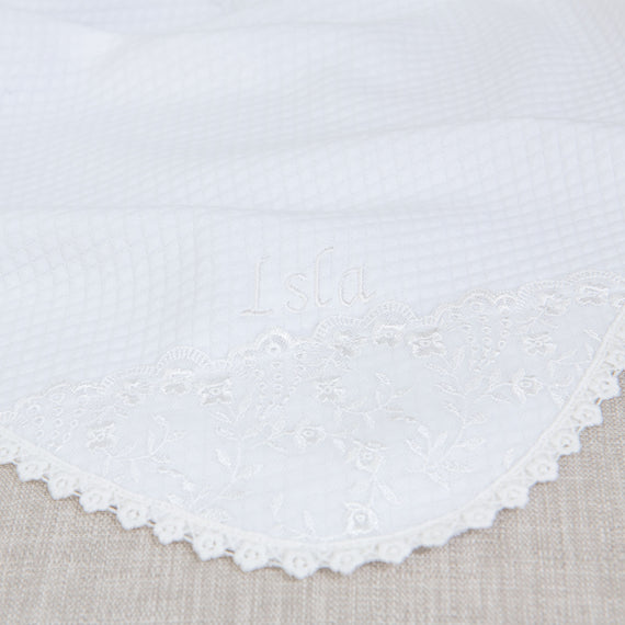 Close-up of the Isla Personalized Blanket, a soft textured white cotton blanket with the name "Isla Personalized Blanket" embroidered on the corner and floral lace border around the edges..