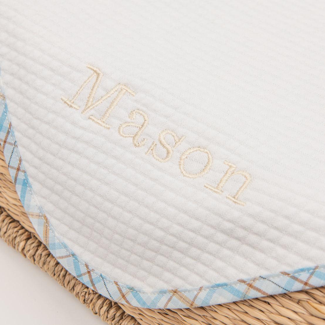 Close-up of a white textured Mason Personalized Blanket embroidered with the name "Mason" in gold for a christening, resting on a woven straw surface with a blue and white checked trim.