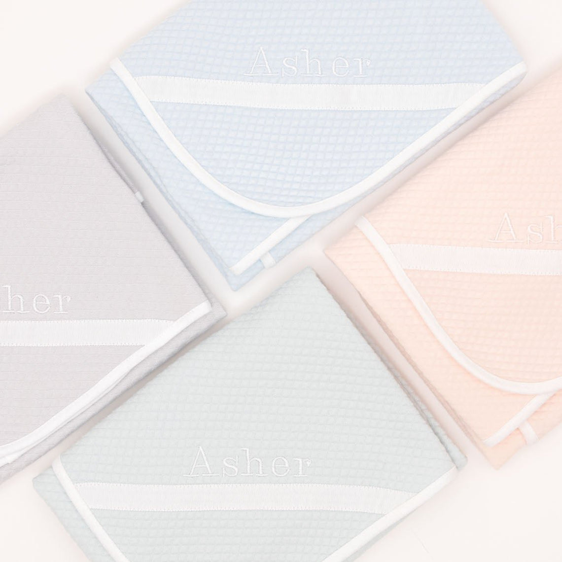 Flat lay photo of the four available colors of the Asher Personalized Blanket (folded in the photo), including grey, pink, powder blue, and soft teal. The blanket is made out of a 100% Quilted Cotton and features an embroidered name on the corner of the blanket.