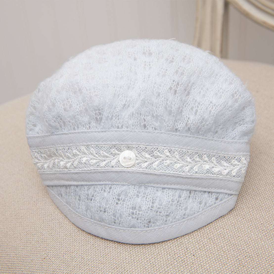 The Harrison Newborn Knit Hat, a light gray knitted beret crafted from the softest knit fabric and decorated with lace and buttons, rests delicately on a beige fabric surface. It's perfect for adding a charming touch to any newborn baby outfit.