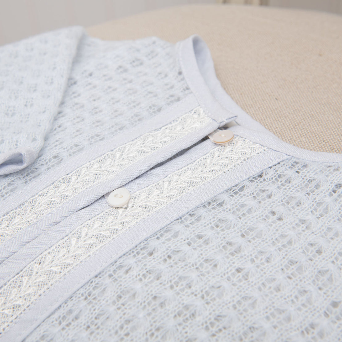 The Harrison Blue Knit Sweater is a handmade baby sweater in a light blue hue, featuring an intricate lace-like design and two white buttons along the center. Captured in a close-up view, the soft knit fabric is laid flat against a textured beige surface.