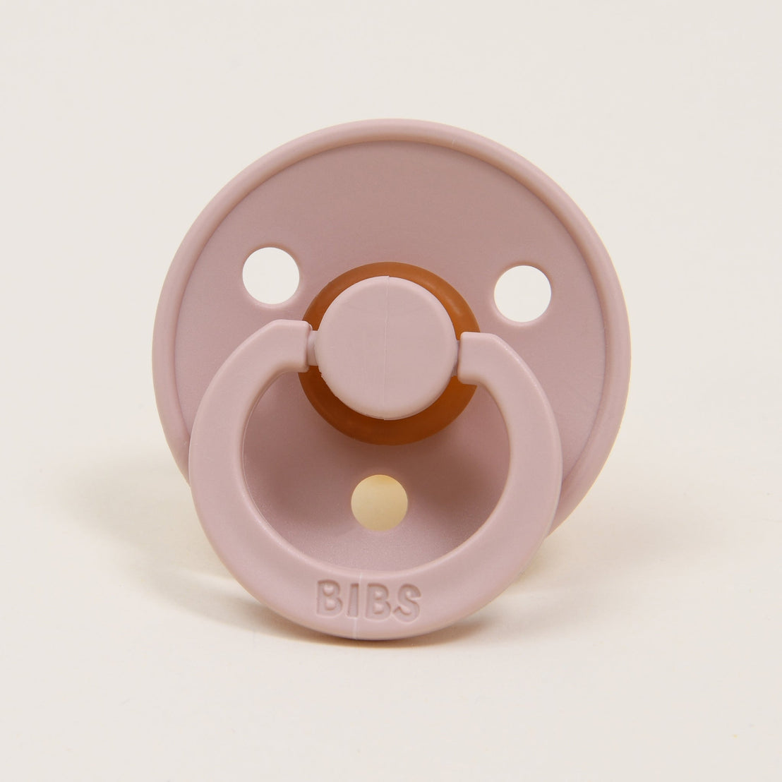 The Tessa Pacifier Set | Ivory & Blush includes a pale pink natural rubber baby pacifier with a round shield that features two large ventilation holes. Designed to aid infants in self-soothing, it comes with a ring handle and has the brand name "BIBS" imprinted on the lower portion of the shield. This pacifier is crafted in Denmark and is set against a neutral off-white background.