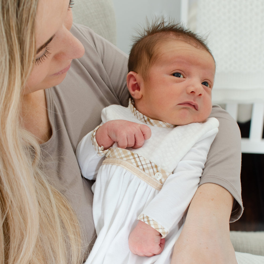 A mother cradles her newborn baby, who is dressed in a white Dylan Layette. They are indoors, and the mother is gazing affectionately at her baby.