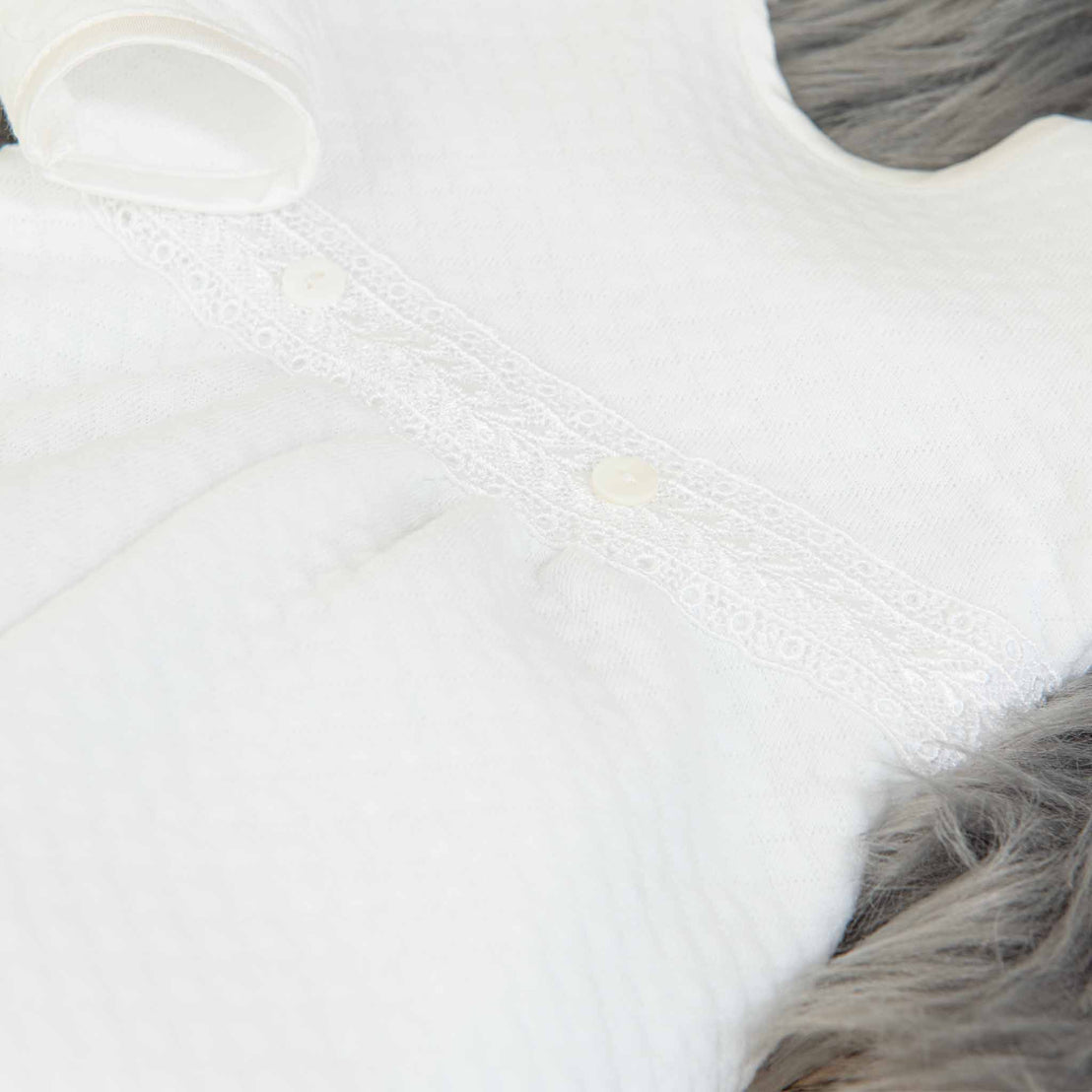 Close-up of a white, textured baby gown from the Elijah Newborn Gift Set - Save 10% with a delicate lace trim and small buttons, lying on a furry gray background. The intricate lace detail includes a subtle pattern, adding elegance to the soft, cozy fabric—perfect as a baby shower gift.