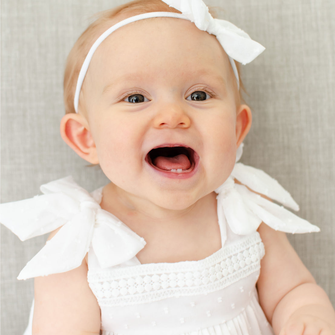 A joyful baby with wide-open eyes and a big smile, dressed in the Mila Cotton Gown and matching Mila Bow Headband sits on a soft gray sofa.