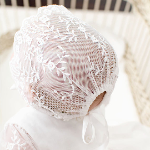 A baby girl inside a crib and wearing an Isla Lace Bonnet viewed from the back. The sheer lace bonnet is made with floral embroidery netting lace with silk ribbon ties.