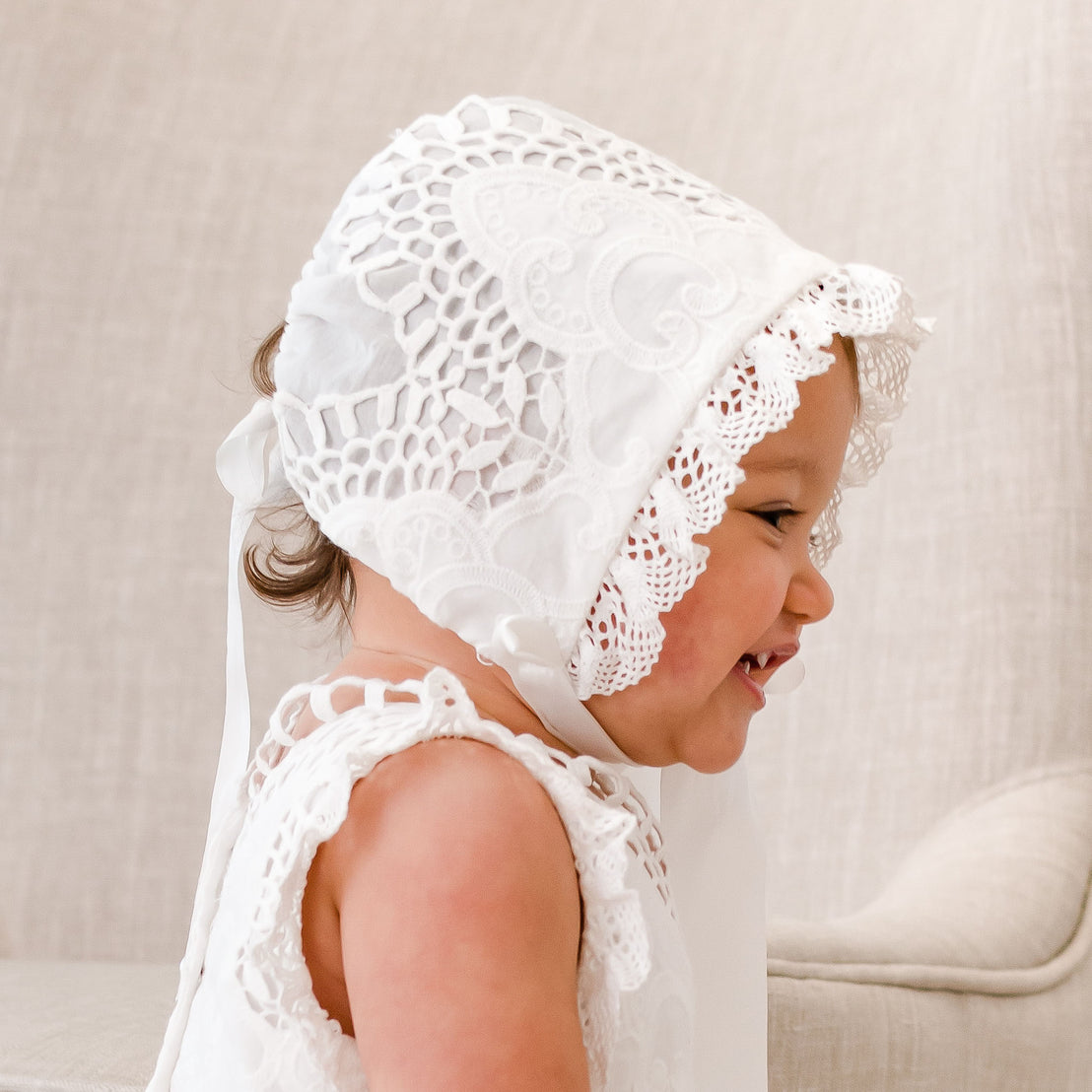 A baby girl wearing a Lily Bonnet looks away from the camera with a grin. The bonnet is made with delicate cotton lace, light ivory edge lace, and ivory silk ribbon.