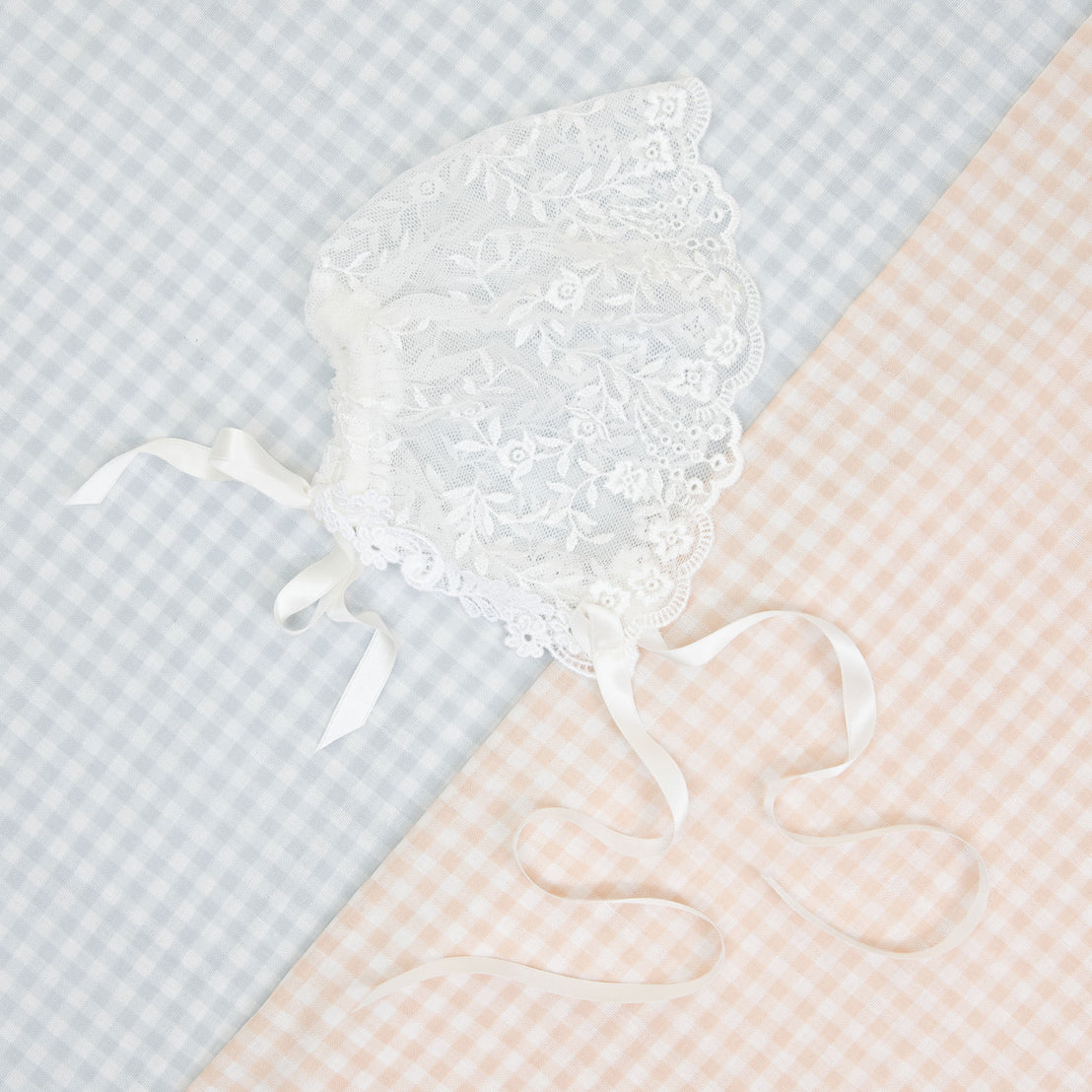 An Isla Lace Bonnet with silk ribbon ties displayed on a pastel blue and peach checkered background.