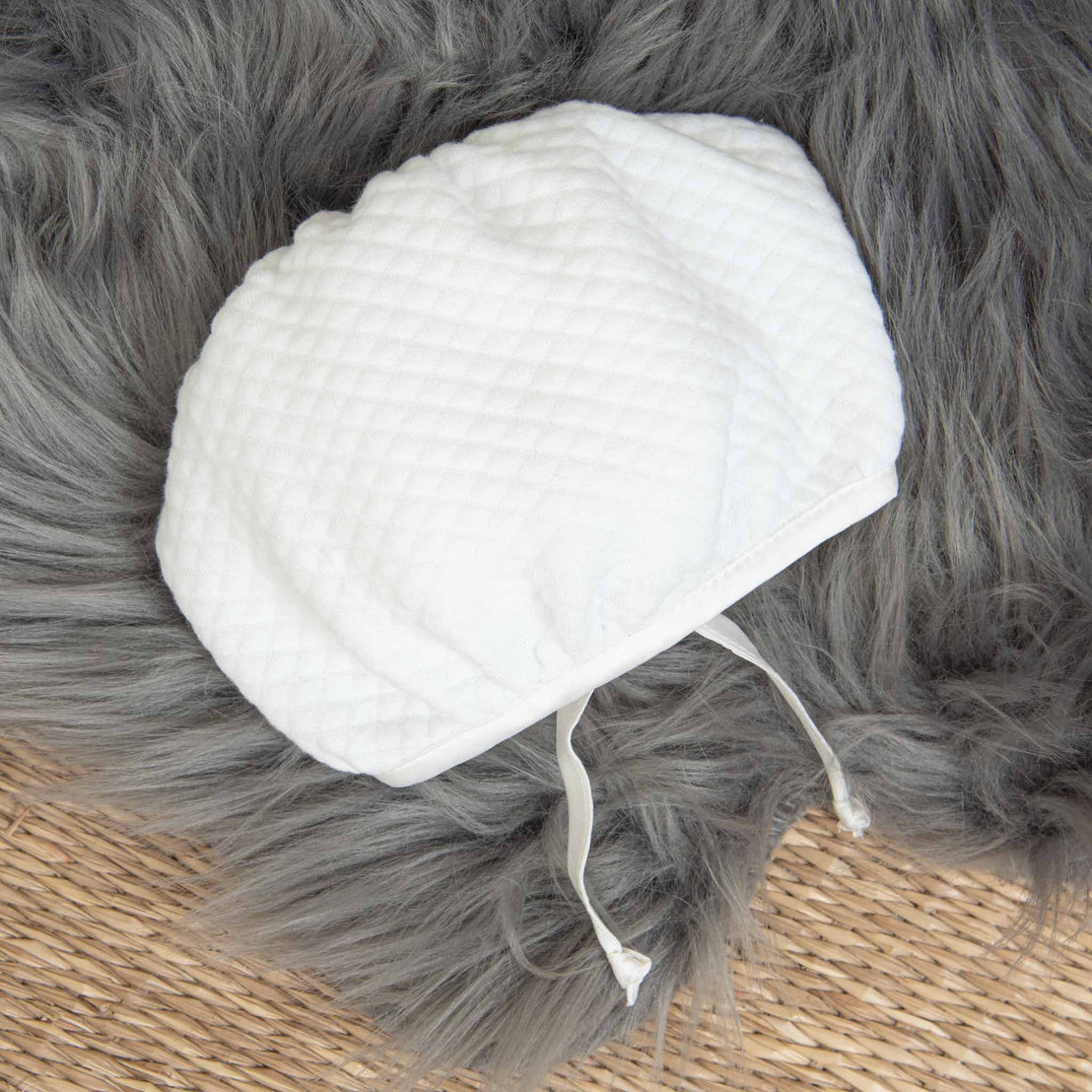 A white, quilted infant bonnet with two ties lies on a fluffy gray surface and a wicker background, part of the charming Elijah Newborn Gift Set - Save 10% that makes for an ideal baby shower gift.