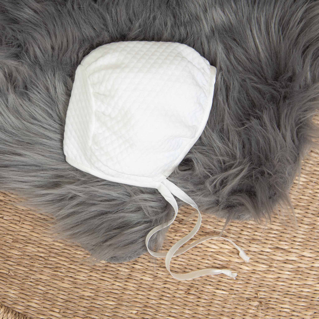 A white quilted baby bonnet with ties is placed on a gray furry fabric and a woven rug background. This soft, simple design is part of the Elijah Newborn Gift Set - Save 10% and showcases its gentle texture, making it an ideal baby shower gift.