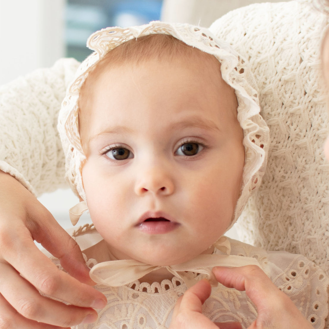 Baby wearing the Ingrid Lace Bonnet crafted with a 100% cotton lace in light ivory and a natural champagne tone with light ivory cotton ribbons 