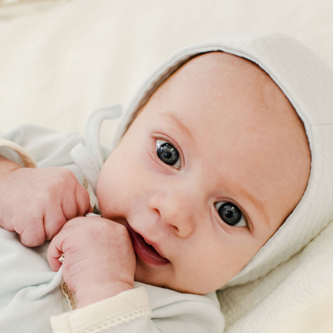 A portrait of a baby with blue eyes and a light complexion, wearing an Aiden Quilt Bonnet and outfit, lying down and looking directly at the camera with a gentle expression.