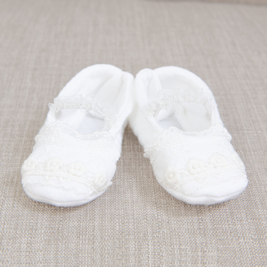 A pair of Isla Booties resting on a textured beige fabric background. The isla booties are crafted with a soft textured cotton in white and detailed with a light ivory floral lace.