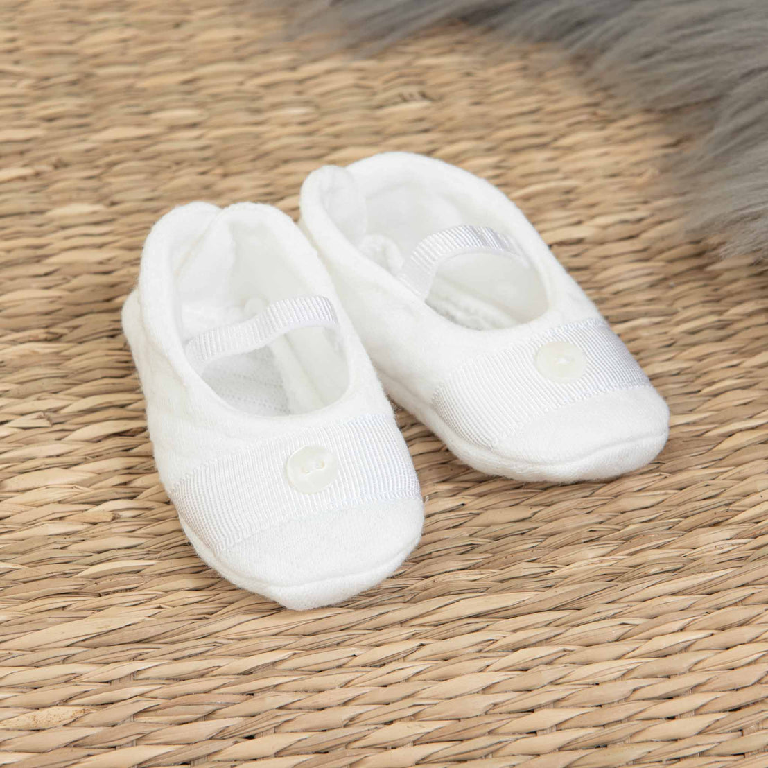 A pair of small, white baby booties set on a woven straw mat. Each bootie features a button detail and a strap over the top. This darling Elijah Newborn Gift Set - Save 10% also includes part of a furry gray blanket in the top right corner, making it an ideal baby shower gift.