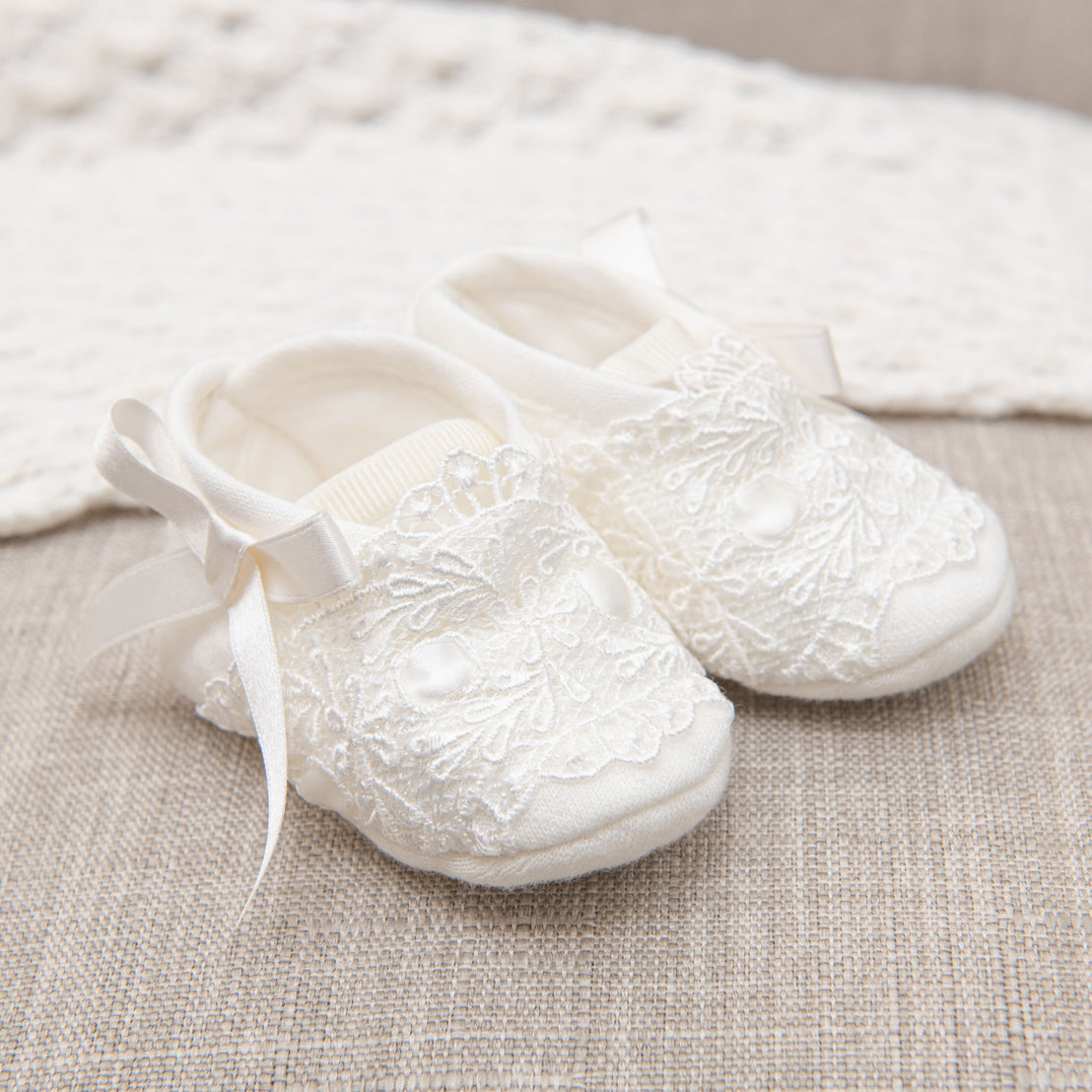 A pair of Madeline Quilted Booties with ribbon ties on a beige fabric background.