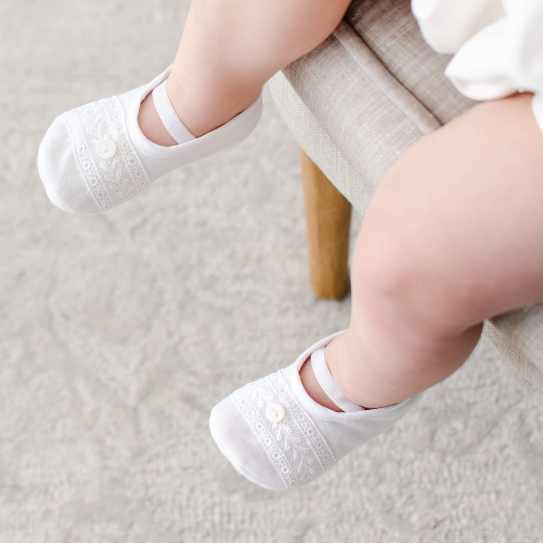 The top detail of the Oliver Booties in white worn by a baby boy. The Oliver booties are made with a linen top, soft pima cotton bottom, and features a Venice lace and button detail (with elastic strap across the foot).