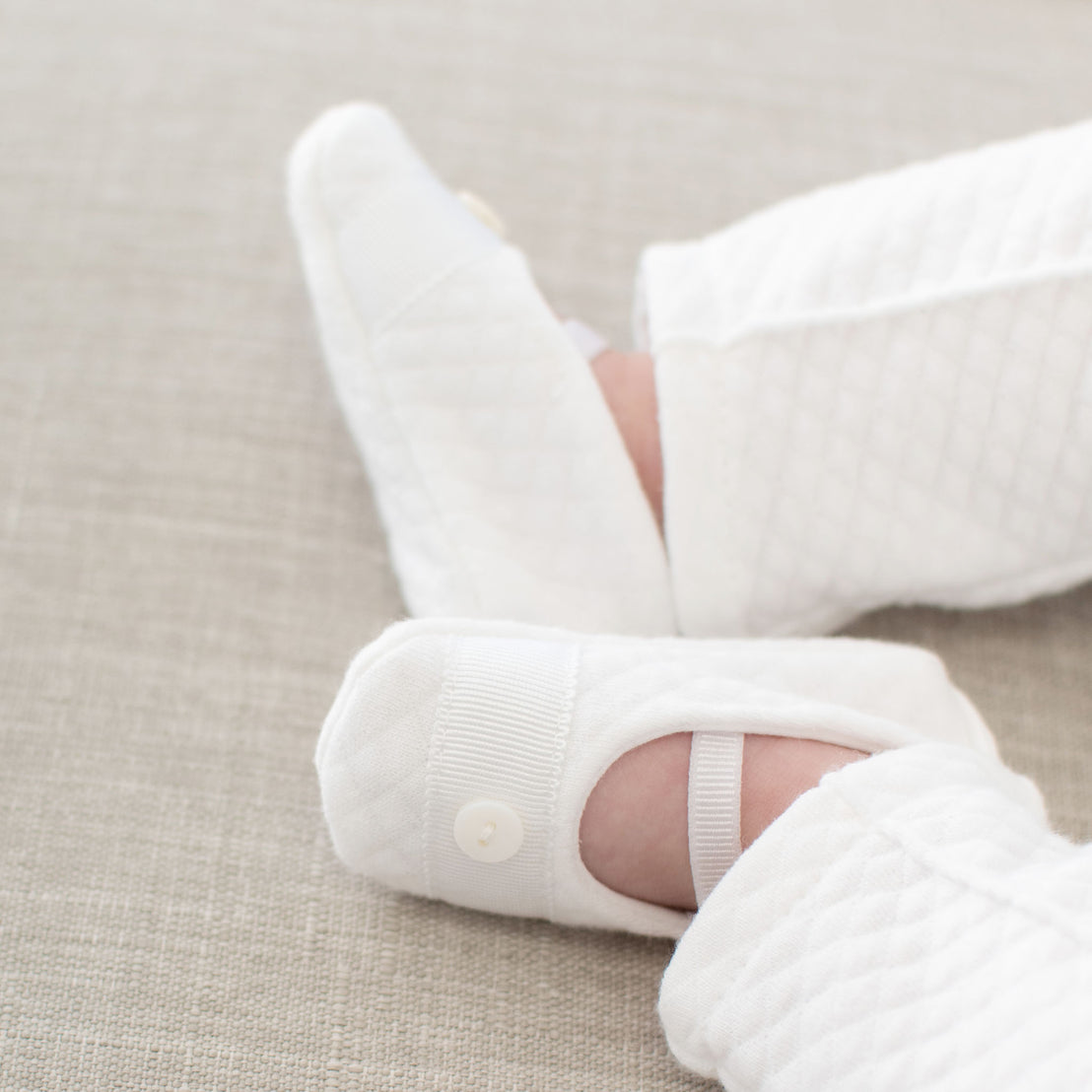 Baby boy wearing the Elijah Booties made from white textured cotton and featuring a grosgrain ribbon trim and button detail across the toe. A soft elastic strap ensures a secure and comfortable fit