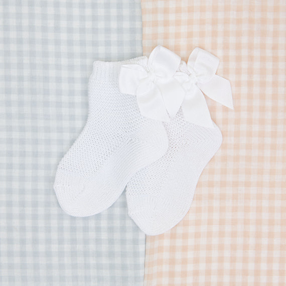 A pair of white newborn Garter Stitch Socks with Bow, displayed on a boutique gingham-patterned fabric background in soft blue and peach tones.