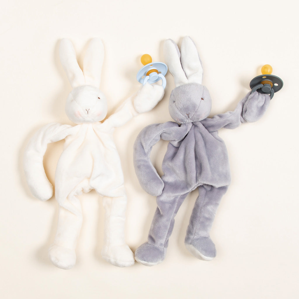 Two Silly Bunny Buddy & Pacifier toys, one white and one gray, designed for newborns, lie side by side with pacifiers attached to their mouths, on a light beige background.